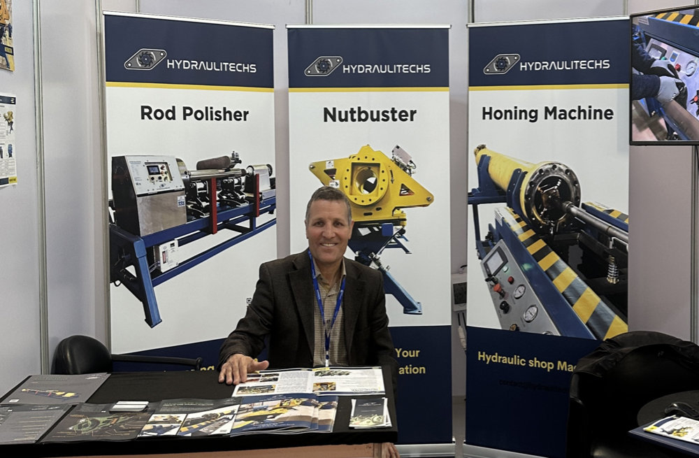 Homme assis à un stand d’exposition pour Hydraulitechs, avec des bannières présentant une polisseuse de tiges, un Nutbuster et une rodeuse